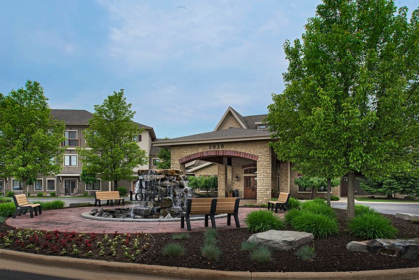 A serene outdoor area featuring a fountain, benches, and lush landscaping in front of a building.