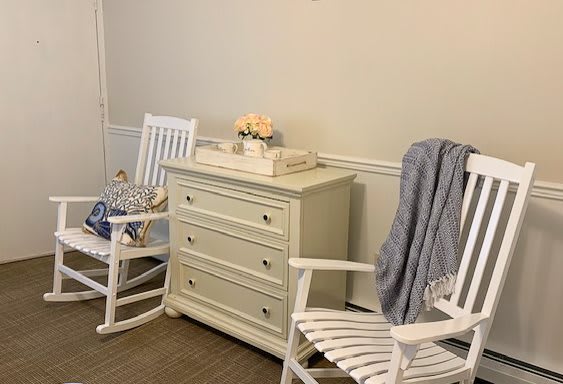 A cozy corner featuring two white rocking chairs and a dresser, decorated with a vase of flowers and a soft blanket.