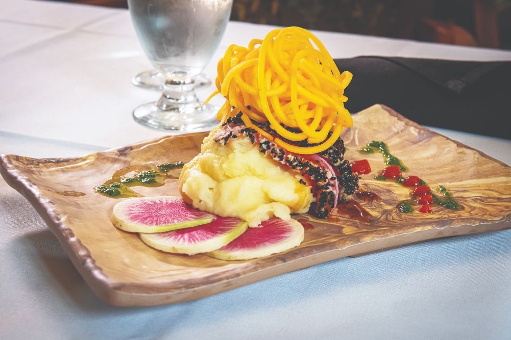 A beautifully presented gourmet dish featuring spiralized garnish atop a main component, with sliced watermelon radish and artistic sauce drizzle on a wooden platter.