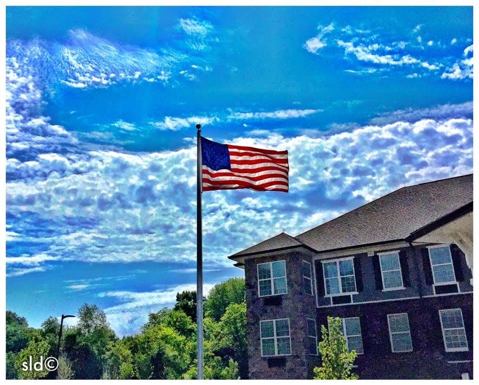 An American flag waves proudly beside a traditional building under a vivid blue sky with scattered clouds.