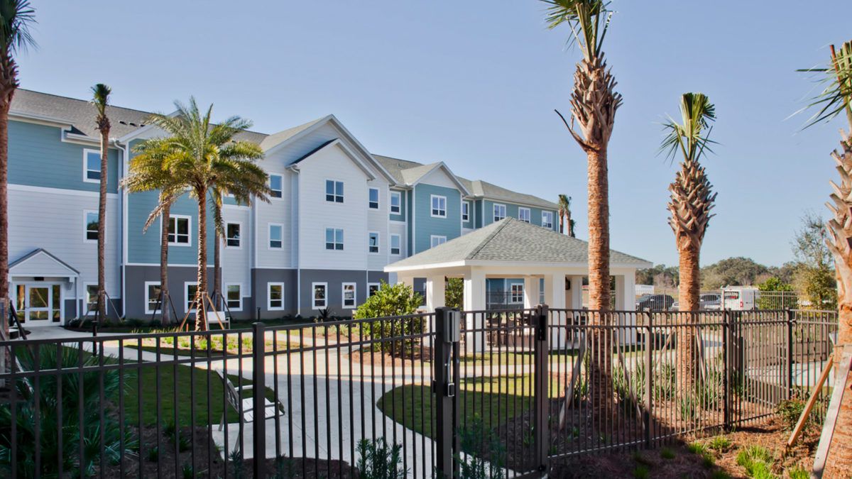 A view of a residential apartment complex with palm trees and a fence in the foreground.