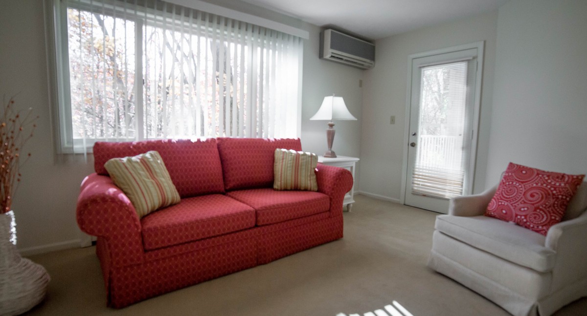 A cozy living room featuring a red sofa adorned with striped cushions, complemented by a white lamp and large windows with sheer curtains.