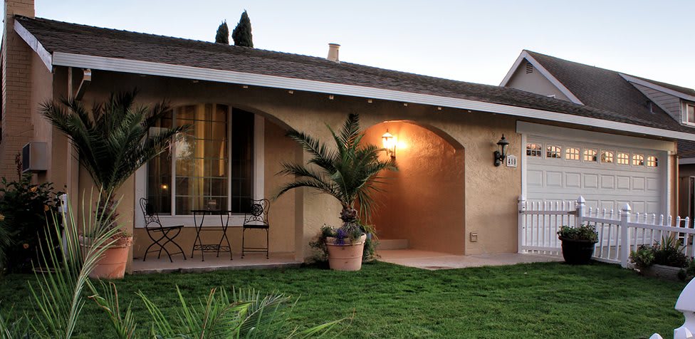 A charming house with a well-lit patio and potted plants in the front yard during the evening.