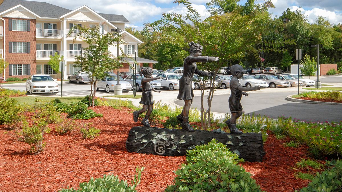 A sculpture of children playing is displayed in an outdoor garden setting with trees and shrubs.