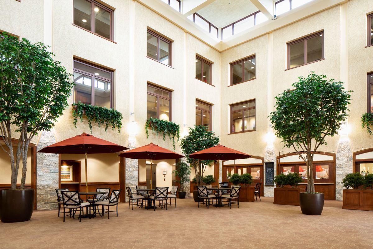 A spacious indoor atrium featuring tables and chairs under red umbrellas, surrounded by greenery and large windows.