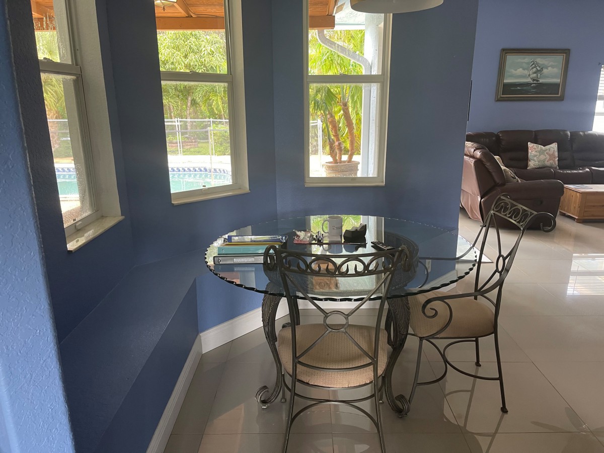 A cozy dining nook with a glass table and chairs, surrounded by blue walls and illuminated by natural light from multiple windows.