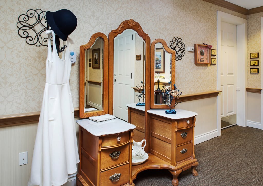 A vintage wooden vanity with a tri-fold mirror, accompanied by a white dress and a black hat hanging on the wall.