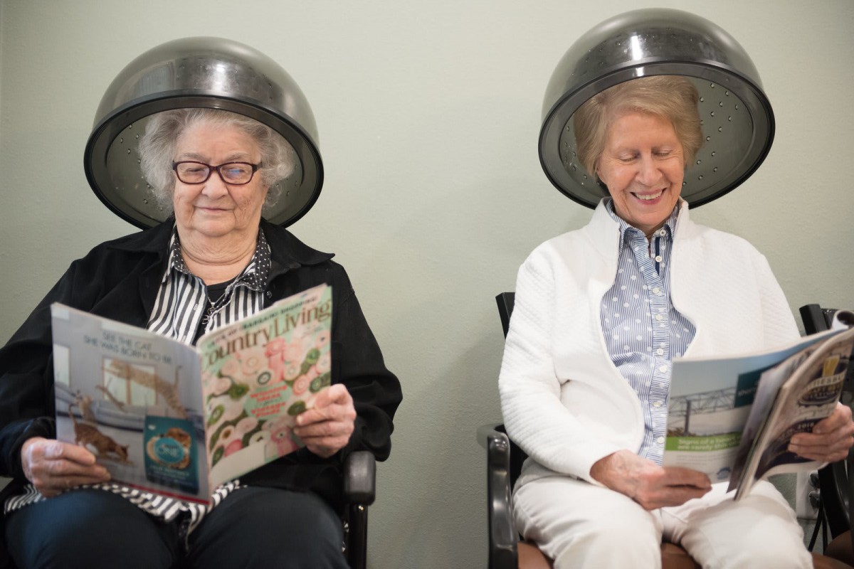 Two older women enjoying a relaxing moment under hair dryers while reading magazines at the salon.