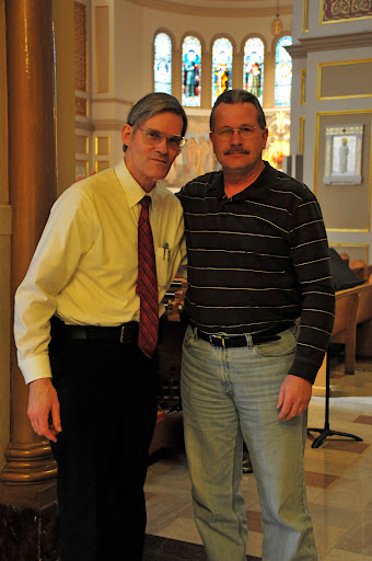 Two men posing together indoors, one in formal wear and the other in casual clothing.