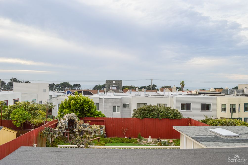 A suburban neighborhood with white houses, a red fence, and lush gardens under a cloudy sky.