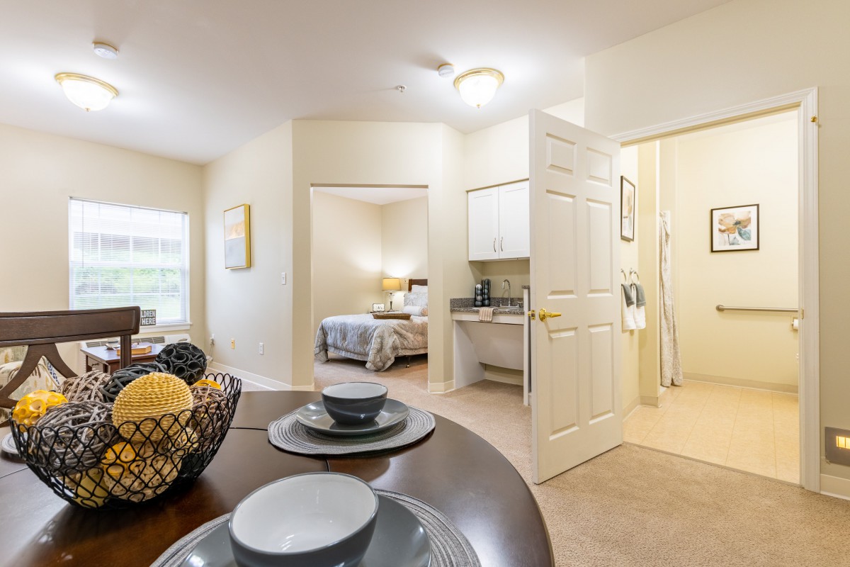 A bright and cozy apartment interior with a dining table set, a view into the bedroom, and a small kitchenette.
