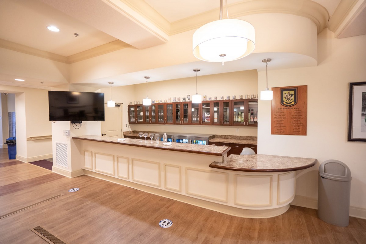 A well-lit bar interior featuring a wooden counter, shelves with bottles, and a mounted television.