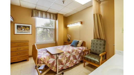 A cozy and well-lit patient room in a hospital, featuring a bed, an armchair, and natural light from the window.