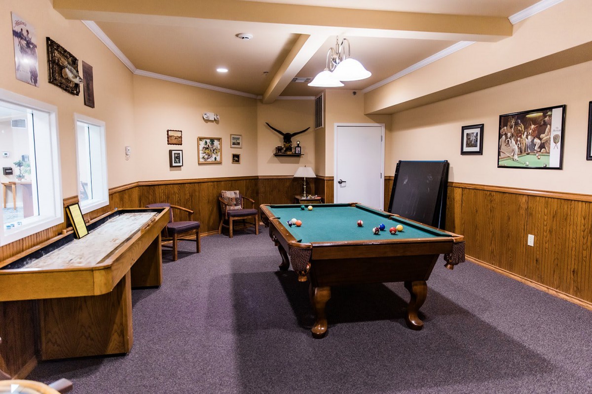 A cozy game room featuring a pool table and a shuffleboard setup, with wooden paneling and framed decor on the walls.