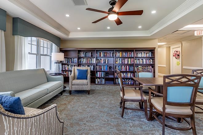 A cozy library room featuring bookshelves, comfortable seating with a sofa and armchair, and a round table under a ceiling fan.