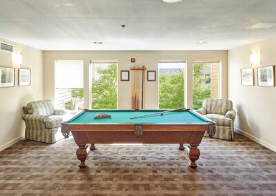 A well-lit game room featuring a pool table surrounded by comfortable seating and large windows.