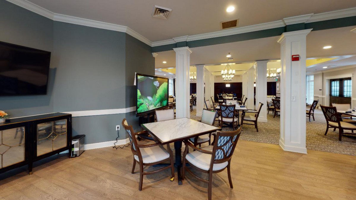 A well-furnished dining area with multiple tables, chairs, and a television, featuring elegant lighting and hardwood floors.