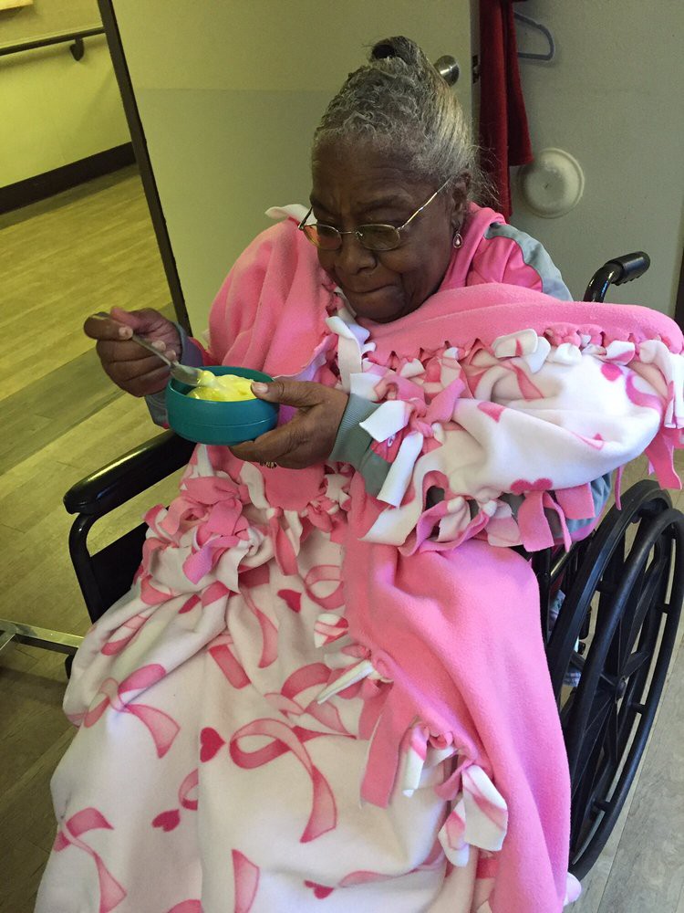 Elderly woman in a wheelchair enjoying a meal, wrapped in a pink blanket.
