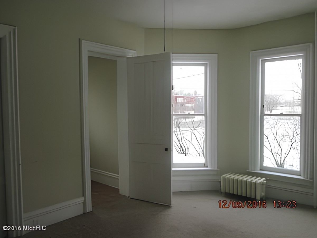 An empty room with two large windows, an open door, and a radiator under one of the windows.
