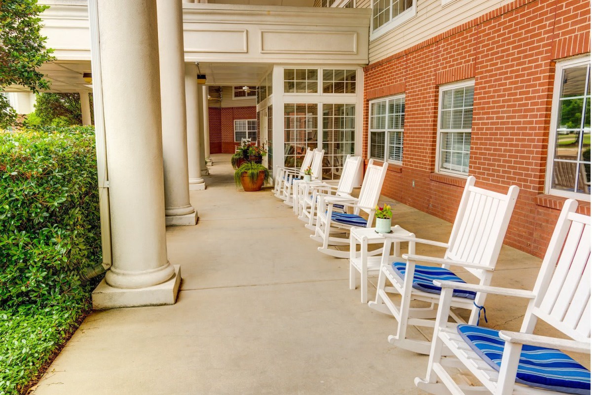 A row of white rocking chairs with blue cushions on a porch outside a brick building.