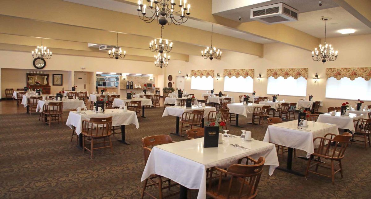 An empty restaurant dining room with white tablecloths and ornate chandeliers.