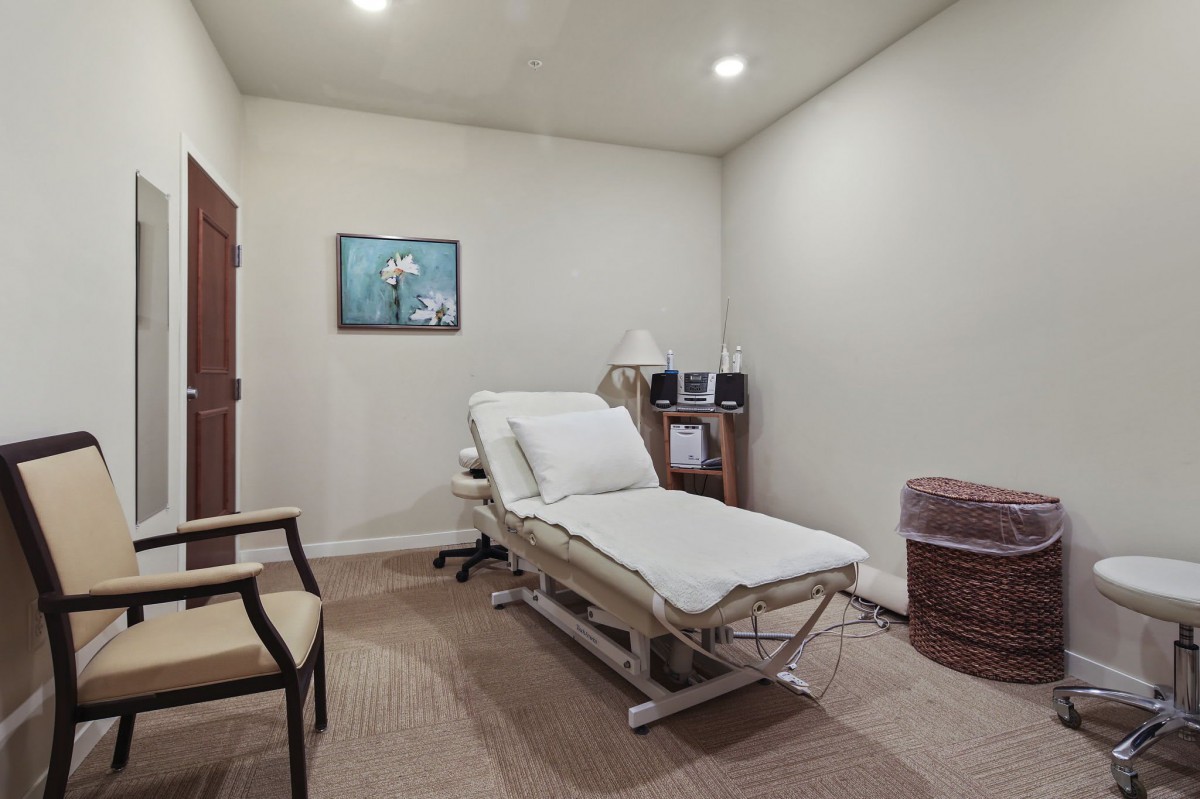 A simple and clean medical examination room with a treatment bed, chair, lamp, and wall art.