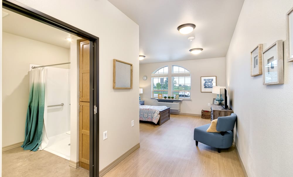 A spacious bedroom with an attached bathroom, featuring a bed, a blue armchair, and large windows.