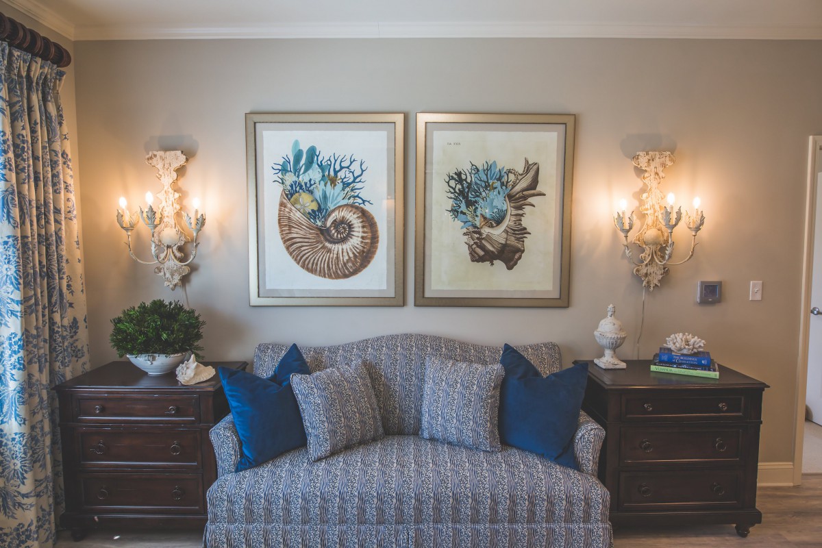 A cozy living room setup featuring a patterned sofa, blue cushions, and sea-themed artwork on the wall, illuminated by elegant sconces.