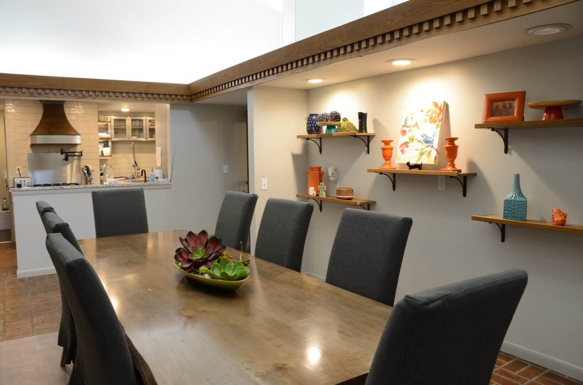A modern dining room with a long wooden table, dark chairs, and decorative shelves displaying artwork and vases.