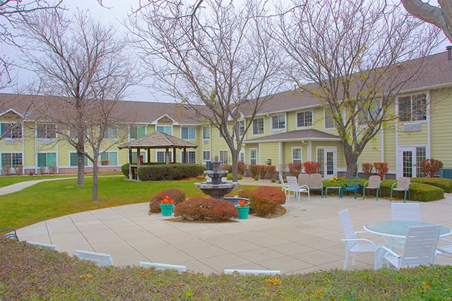An outdoor patio area with a central fountain, surrounded by trees and benches, adjacent to a two-story building.