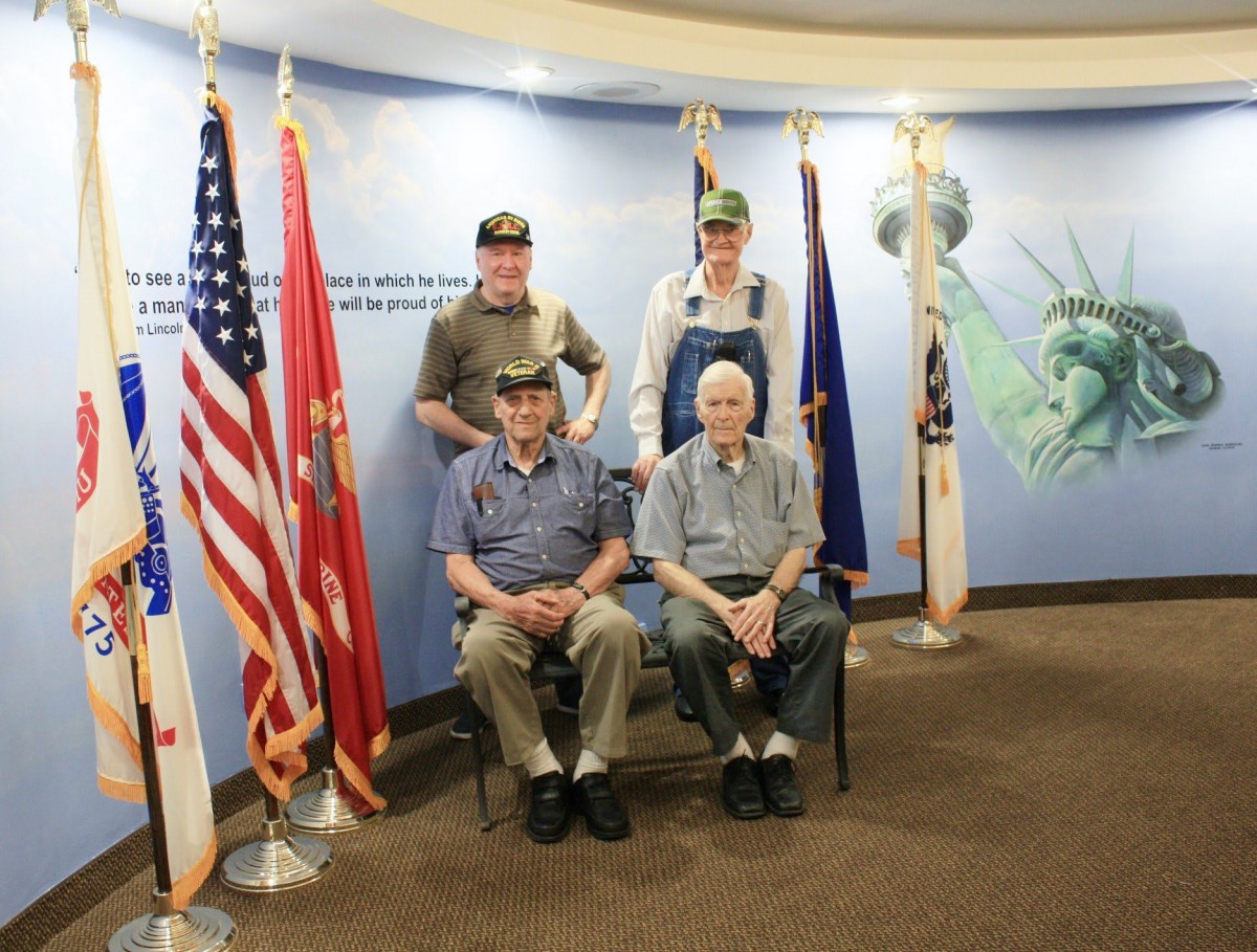 A group of veterans pose for a photo with military flags in the background.