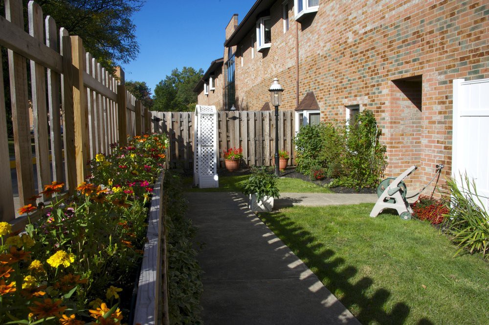 A garden with a pathway, bordered by flowers and a wooden fence, next to a brick building.