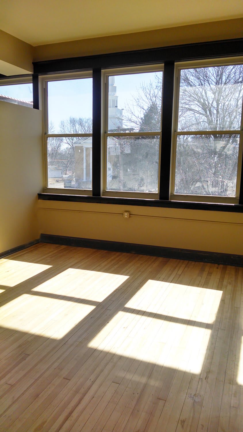 An empty room with large windows allowing sunlight to stream onto the hardwood floor.