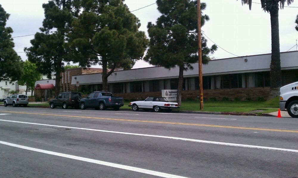A street view showing parked cars alongside a sidewalk with trees and a building in the background.