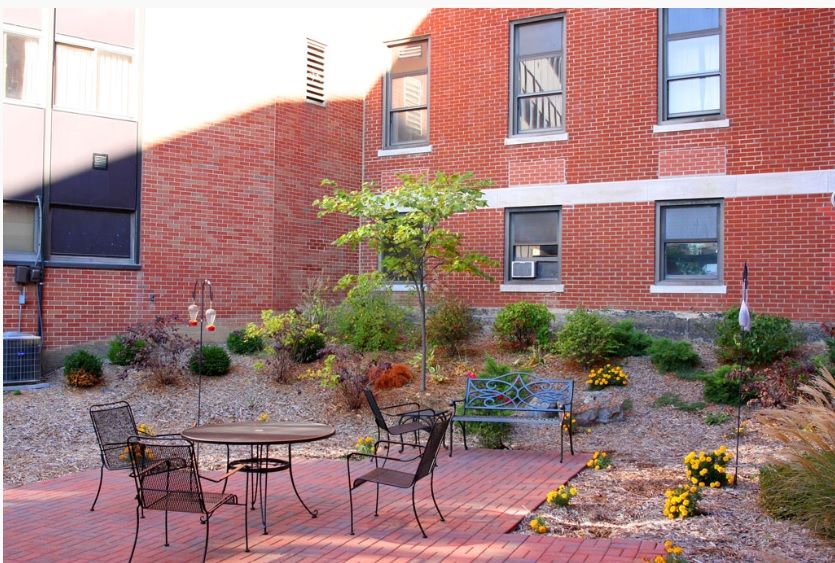 A quaint outdoor courtyard with tables and chairs, surrounded by a landscaped garden adjacent to a brick building.