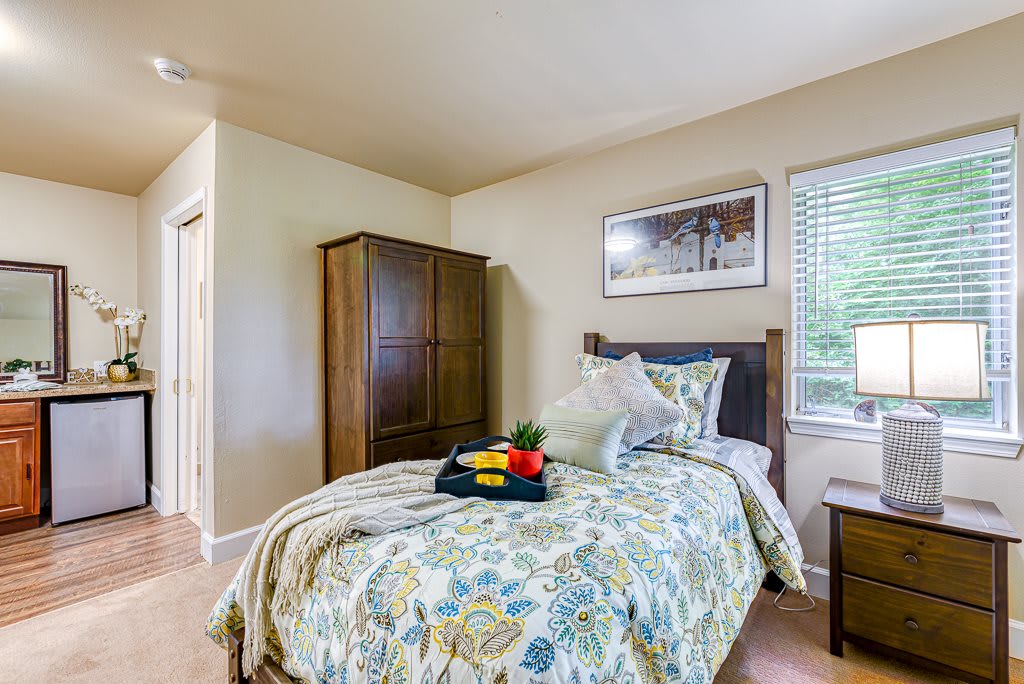 A cozy bedroom with a single bed dressed in floral bedding, featuring a wooden wardrobe and a window that lets in natural light.