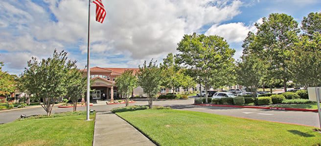 A building with a parking lot, surrounded by trees and under a cloudy sky, with a flagpole in front.
