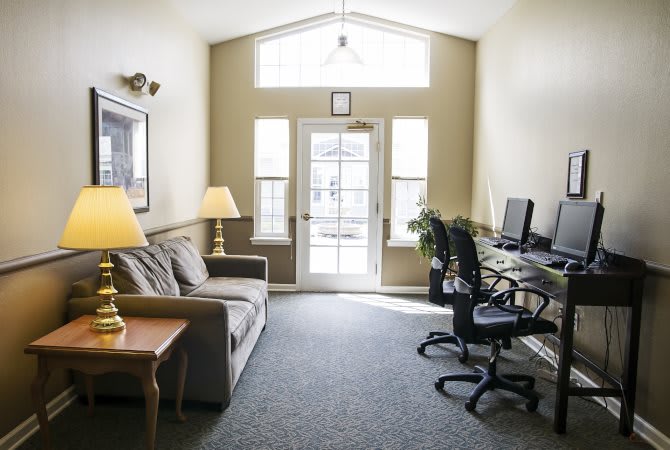 A bright and cozy multi-purpose room featuring a sofa, two computer stations, and large windows.