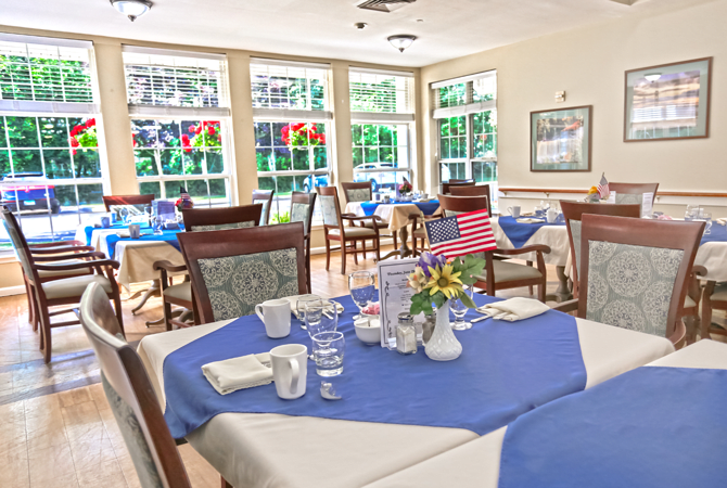 A bright and inviting dining room with tables set with blue napkins and white tablecloths, featuring decorative stained glass windows.