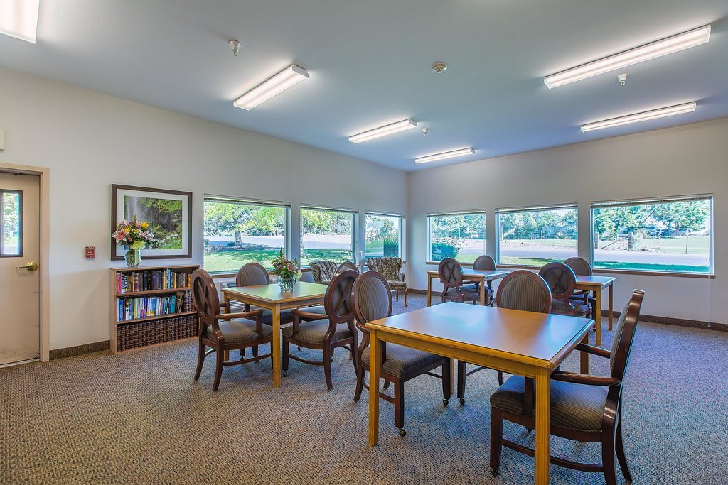 A quiet library room with tables and chairs, surrounded by bookshelves and large windows.