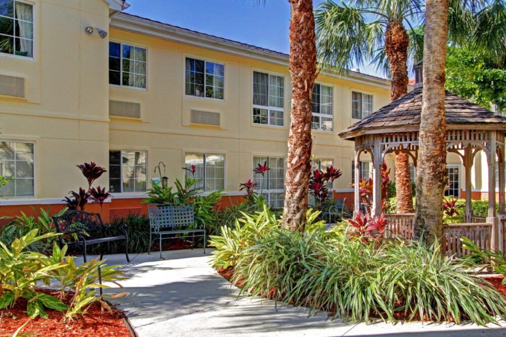 A sunny outdoor garden area with palm trees and a gazebo in front of a two-story building.