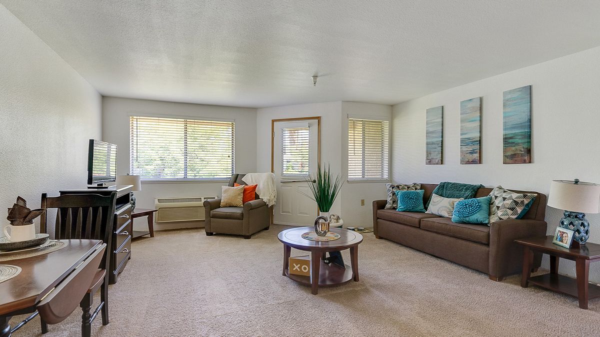 A spacious living room with neutral tones, featuring a sofa, armchair, and a central coffee table, with ample natural light coming through large windows.