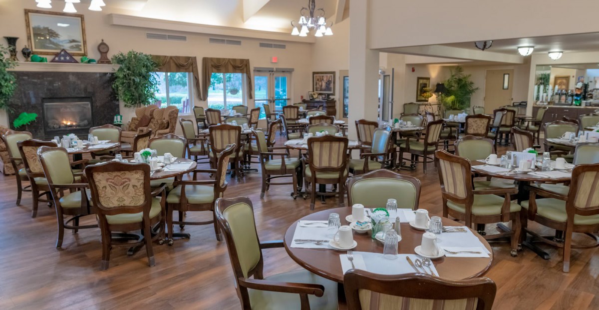 A spacious dining room with elegantly set tables and chairs, featuring large windows and chandeliers.