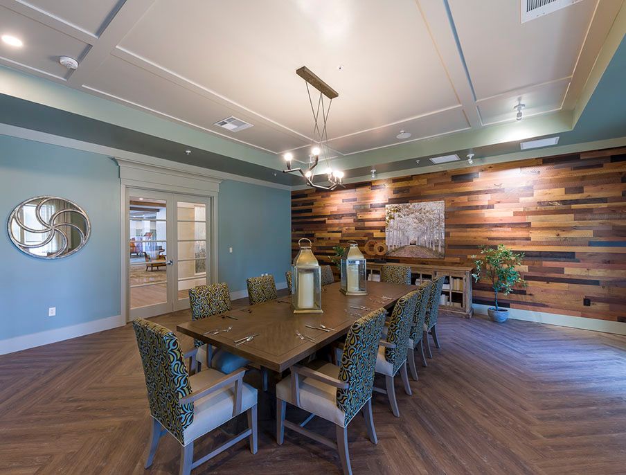 A modern dining room featuring a long wooden table, surrounded by chairs with cushions, and an accent wall of wood paneling highlighted by a chandelier.