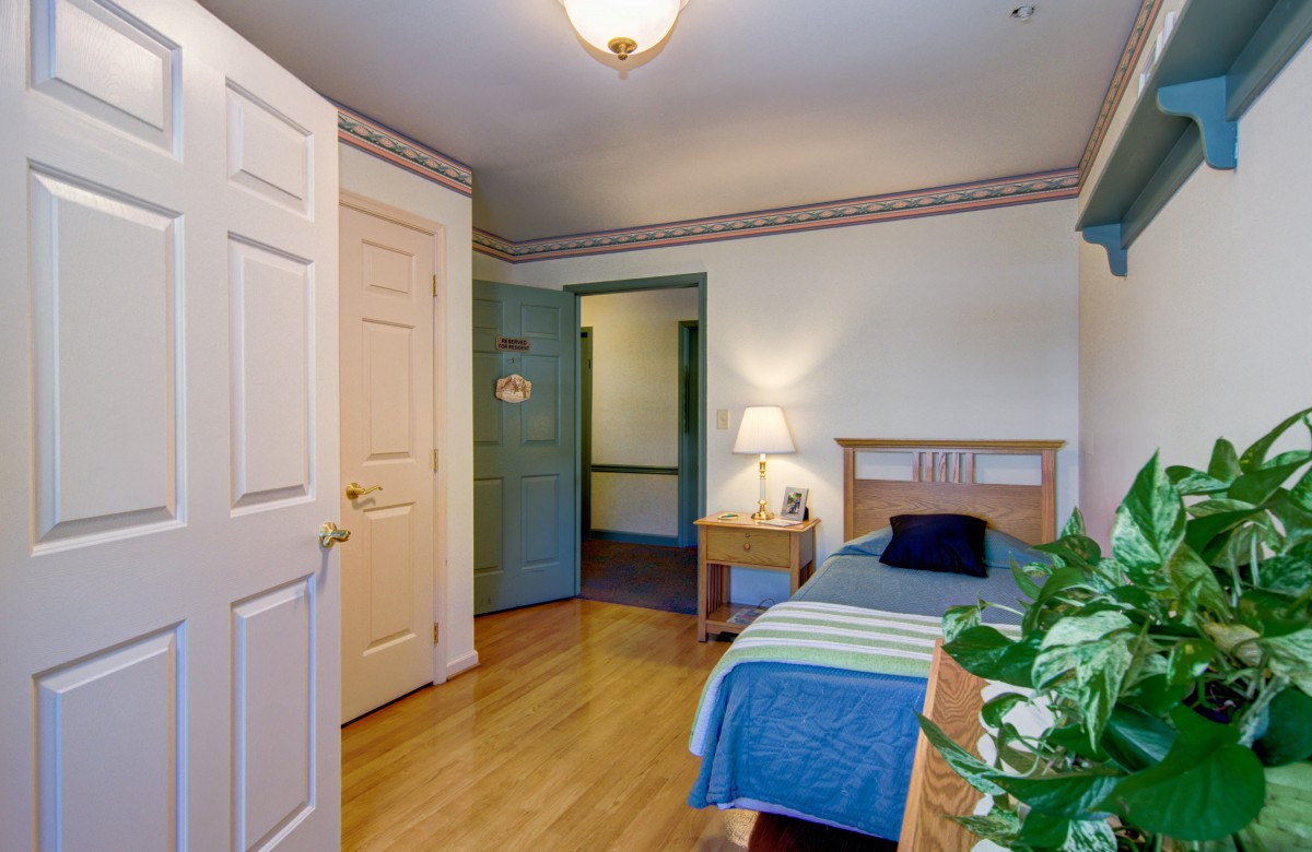 A cozy bedroom with a single bed, nightstand, and lamp, featuring wooden flooring and decorative elements.
