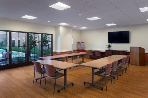 A modern conference room with U-shaped tables and chairs, featuring a large television and glass doors.