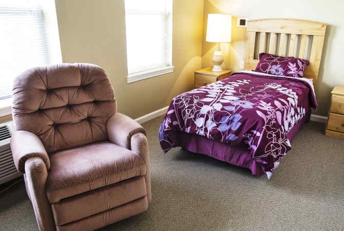A cozy bedroom with a single bed, a comfortable armchair, and a bedside lamp.
