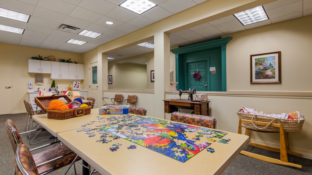 A game room featuring a table with a colorful puzzle in progress, surrounded by chairs and lit by overhead ceiling lights.
