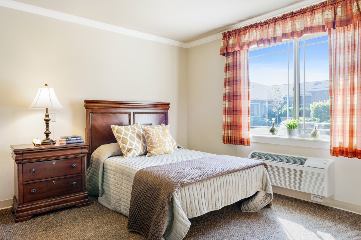 A cozy bedroom with a neatly made bed, wooden headboard, and a large window with plaid curtains.