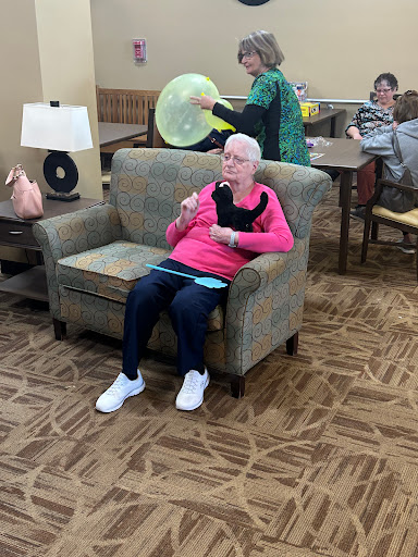 A senior woman enjoying a treat in a community lounge, surrounded by others engaging in various activities.